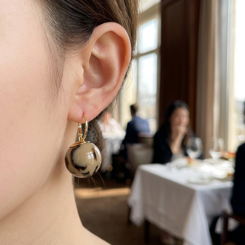 Kolczyk ze złotym zapięciem i kulką khaki z ciemnymi plamami na modelce w restauracji